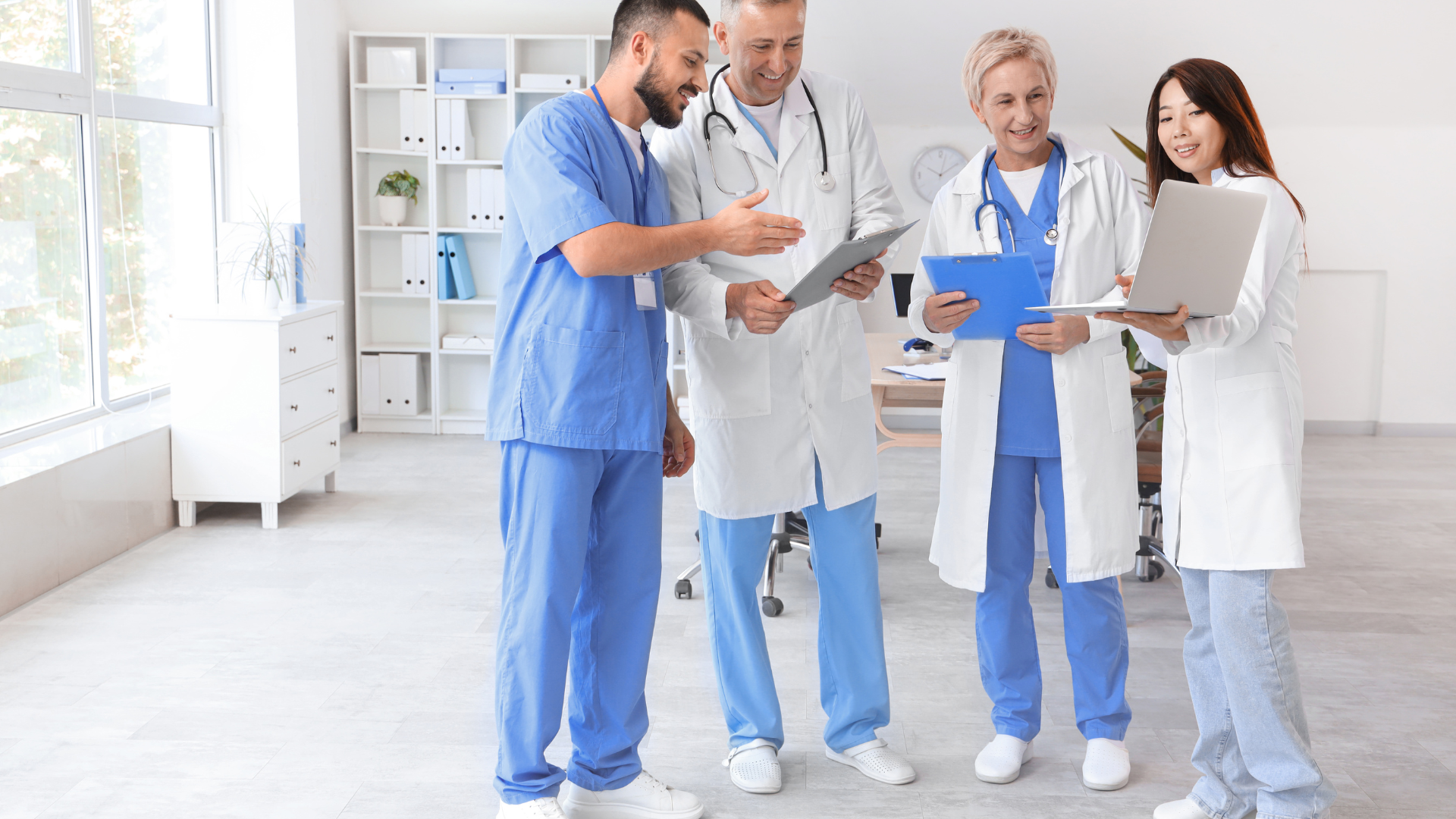 A group of four medical professionals, including doctors and nurses, are gathered in a bright, spacious office. They are discussing a clipboard and laptop, smiling and collaborating with one another in a relaxed setting.