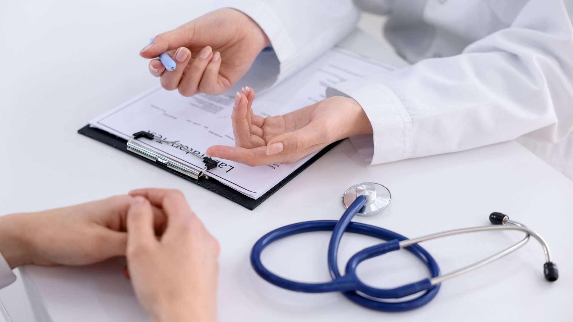 A doctor and a patient are seated across from each other in a clinical setting. The doctor is holding a pen and explaining something to the patient while discussing paperwork. A stethoscope and patient documents are visible on the table.