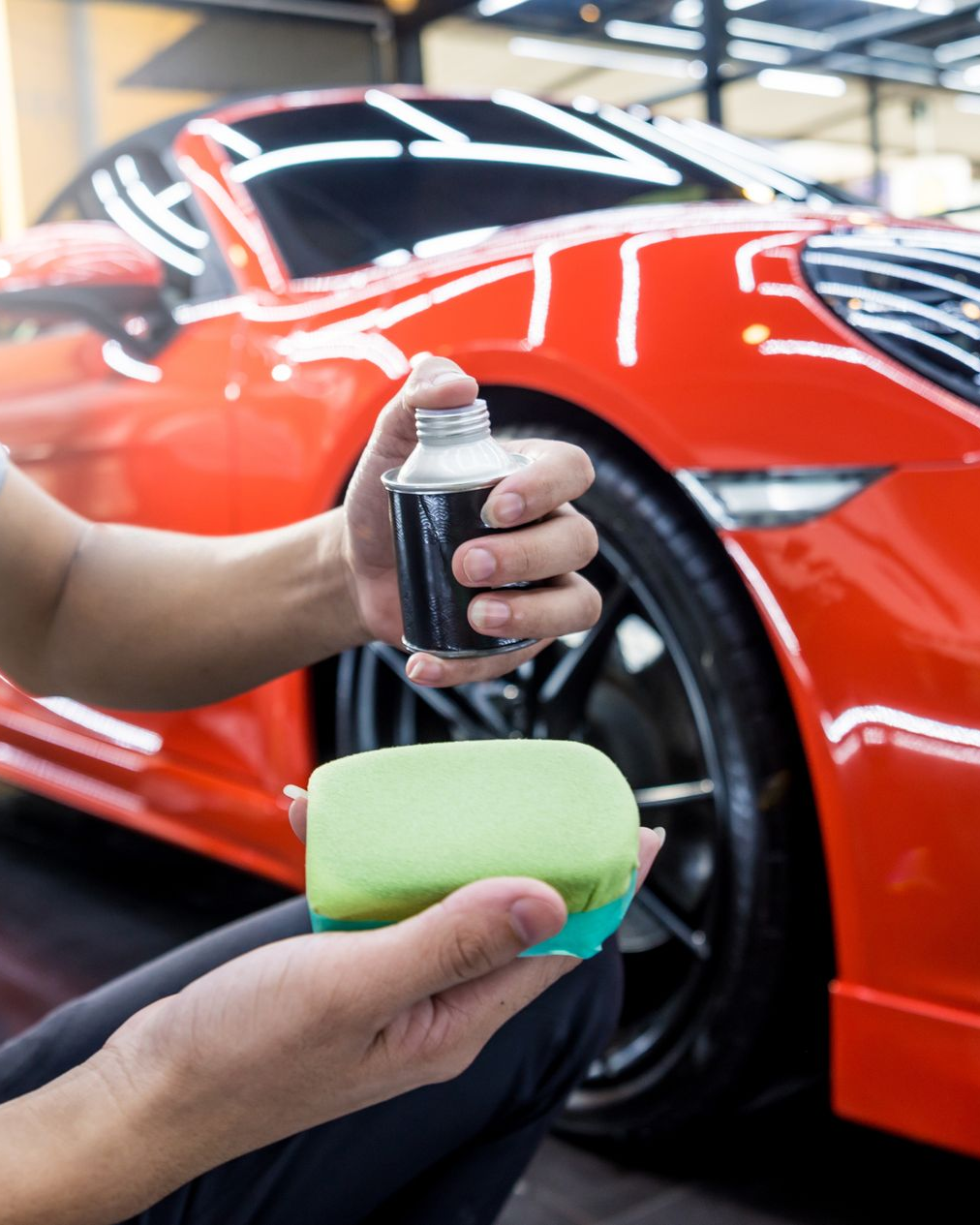 Person applying polish to a red sports car with a sponge in a garage.