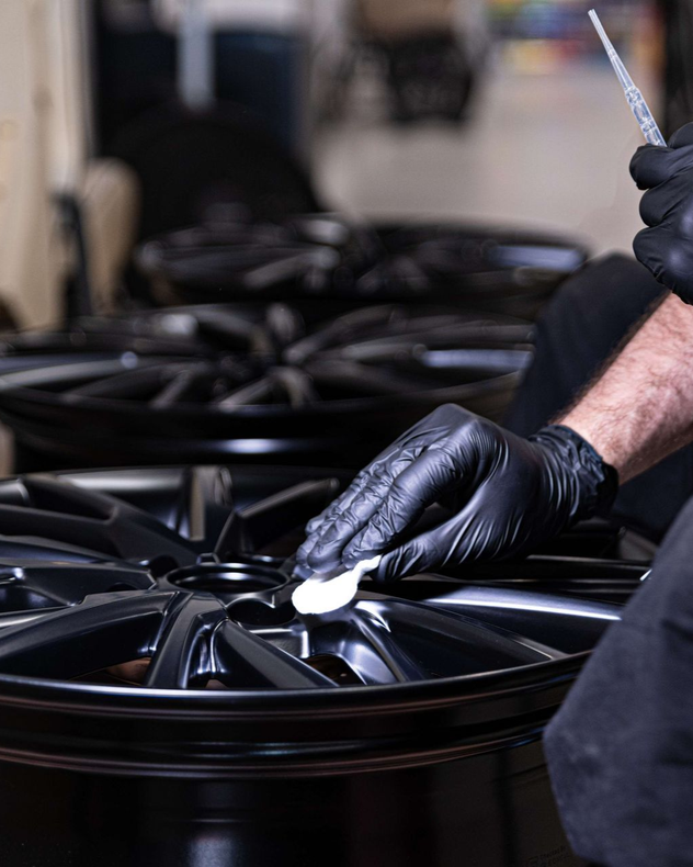 Person wearing black gloves cleaning a black car wheel in a workshop.