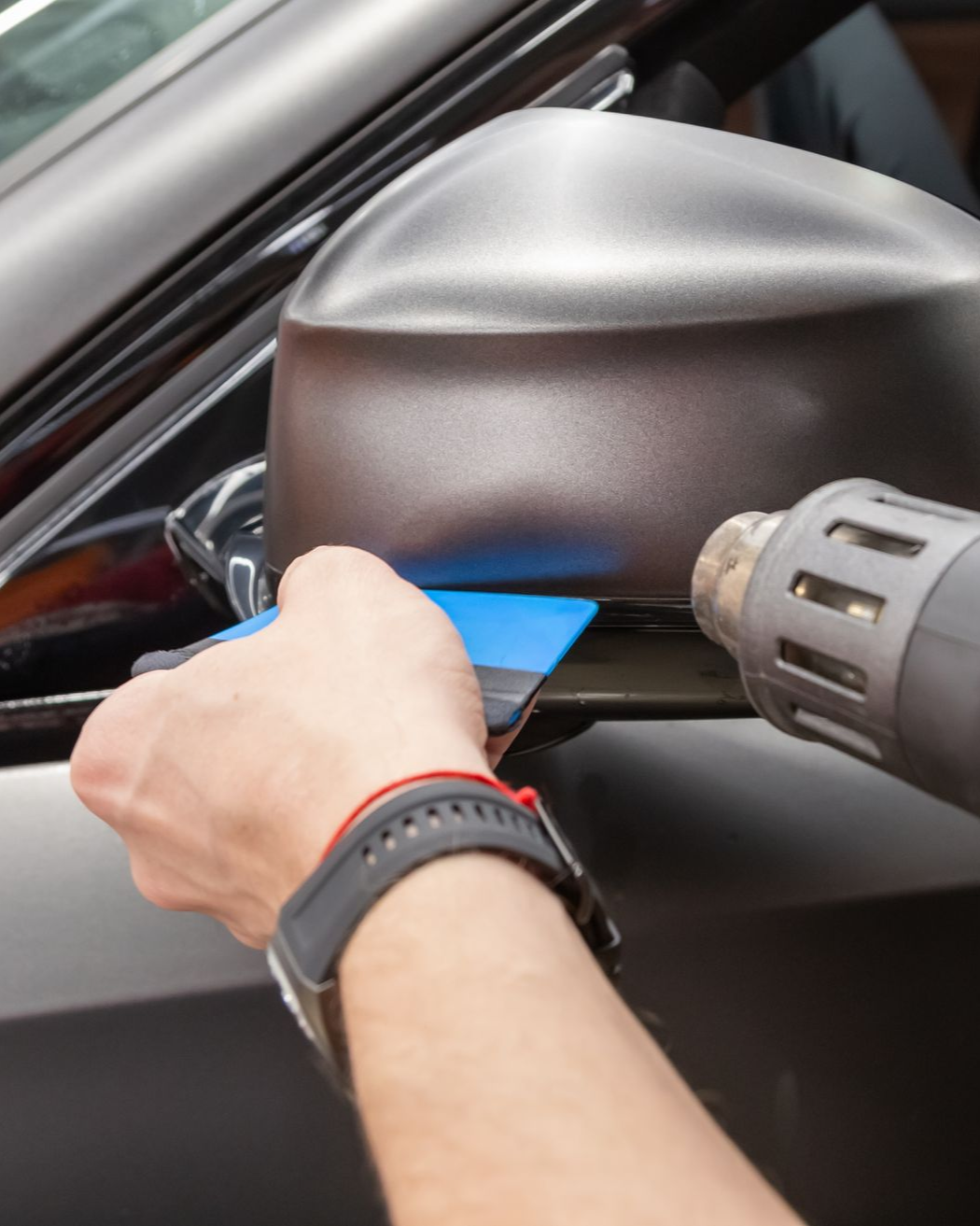 Person using a squeegee and heat gun on a car side mirror wrapped in matte film.