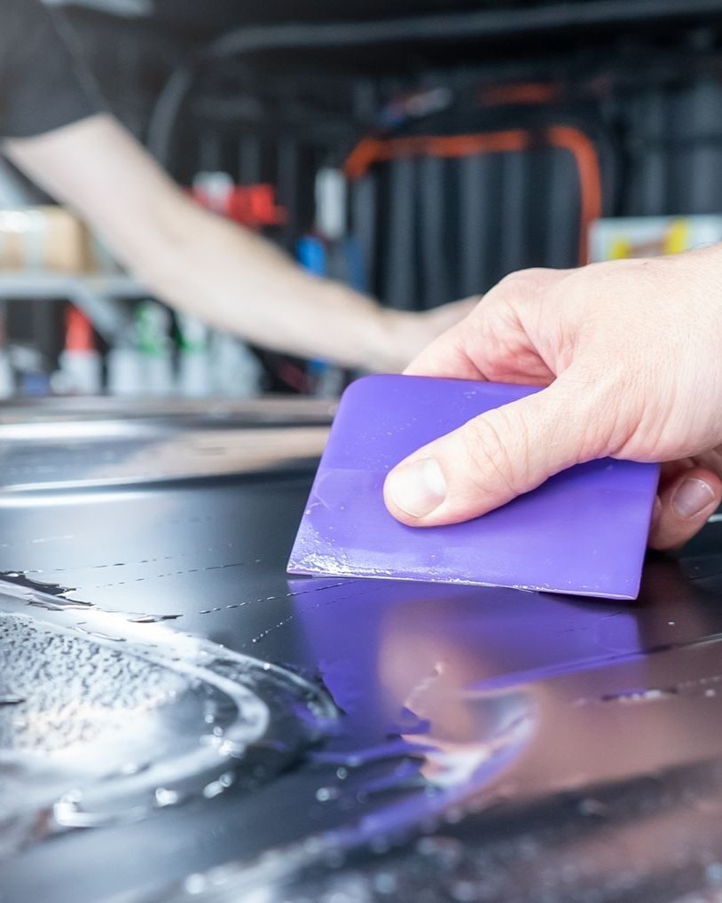 Person using a purple squeegee on a car's surface. Another person is in the background.