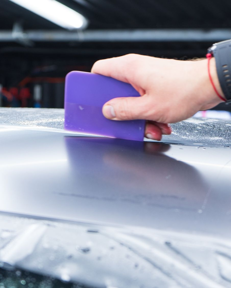 Person using a purple squeegee to apply film to a car's surface.