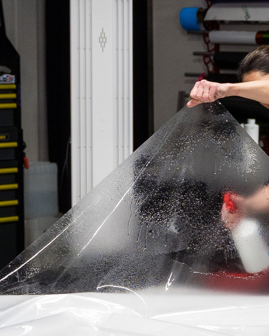 Person spraying water on a large sheet of black film on a white surface, likely applying tint to a car window.