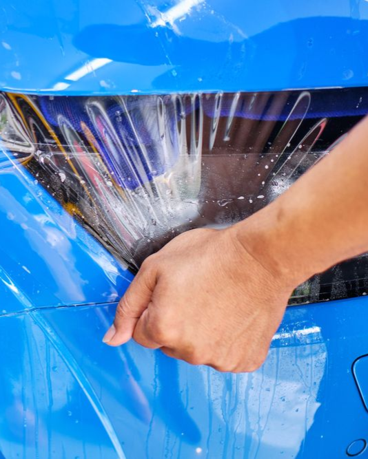 A person is using a blue squeegee to remove plastic from the hood of a car