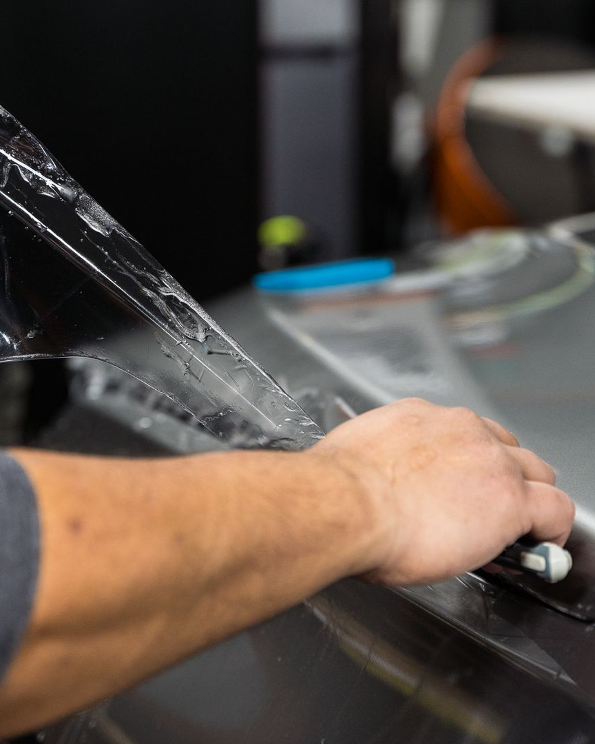 A person is using a blue squeegee to remove plastic from the hood of a car