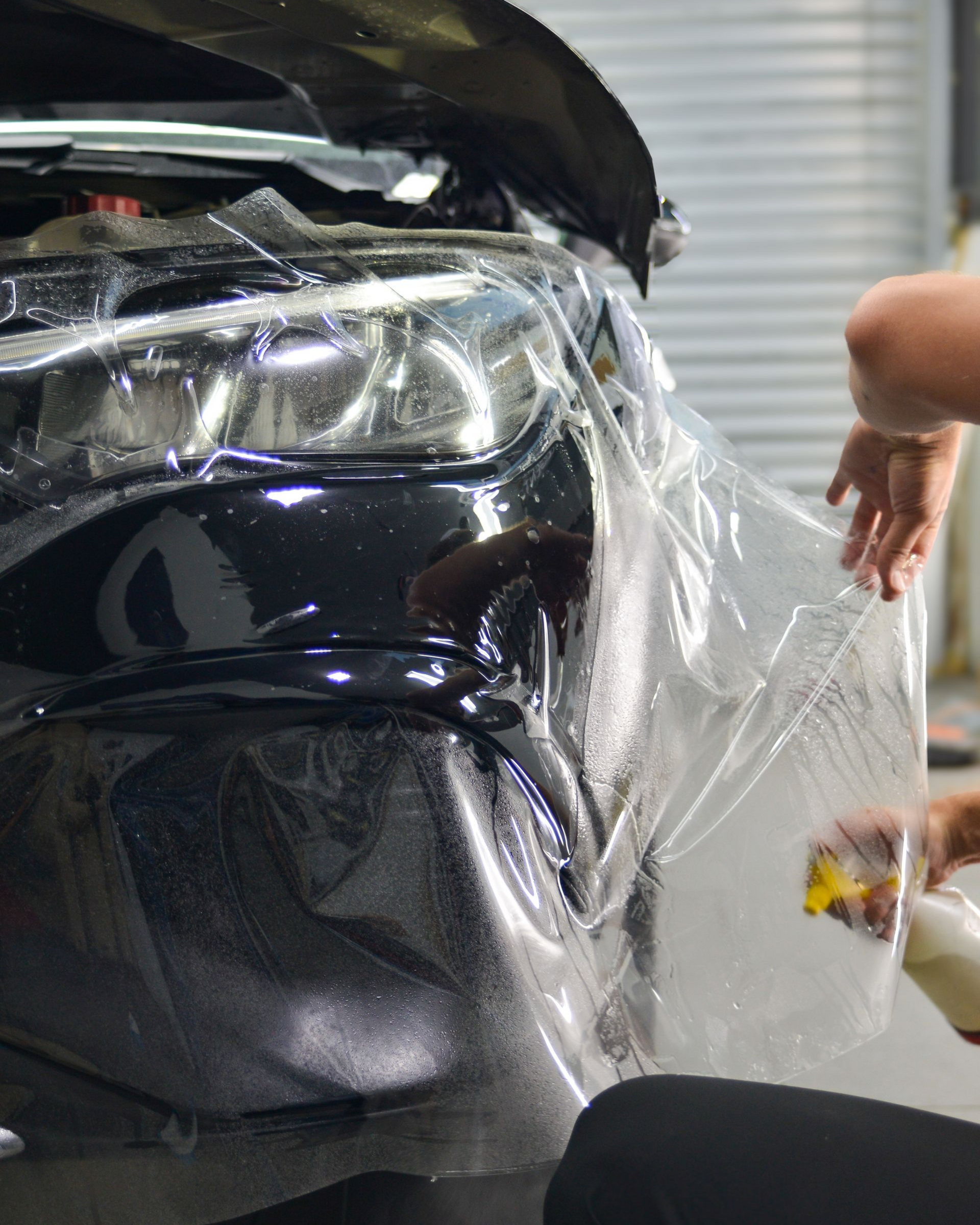 Clear film being applied to a black car bumper and headlight in a garage.
