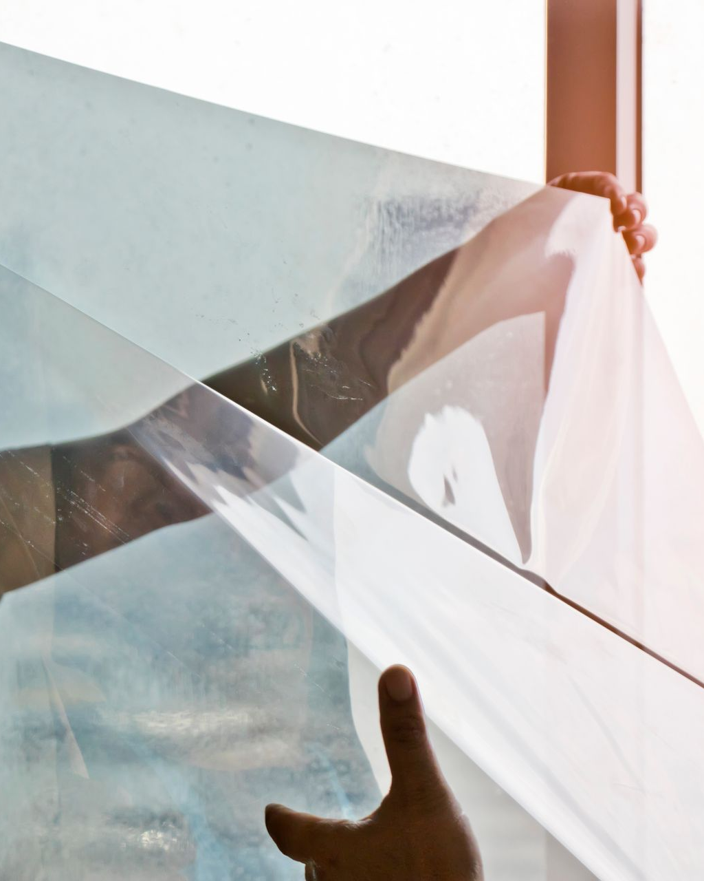 Hands holding and applying a light blue, transparent film to a window, likely for privacy or sun protection.