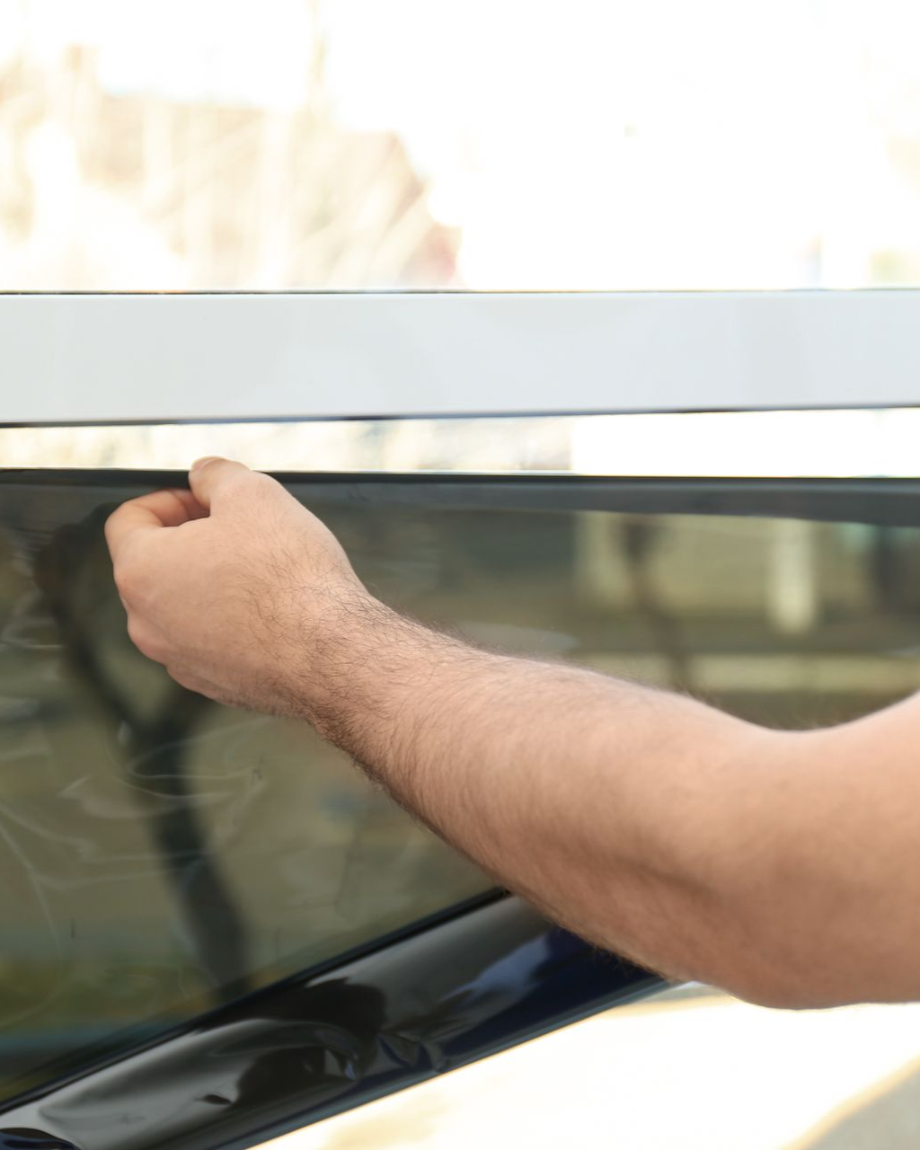 Person's hand applying tinted film to a window, likely for a vehicle, indoors.