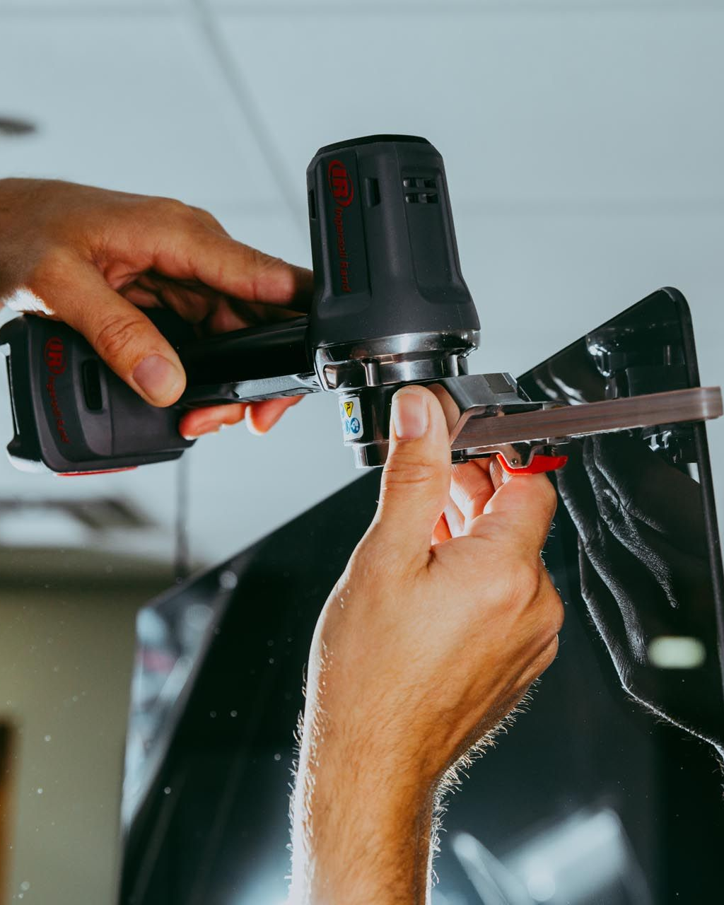 Hands using a handheld tool to trim a dark sheet of material on a car.