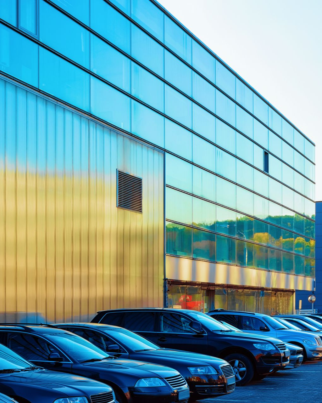Modern building with blue glass facade, parking lot with cars in front.