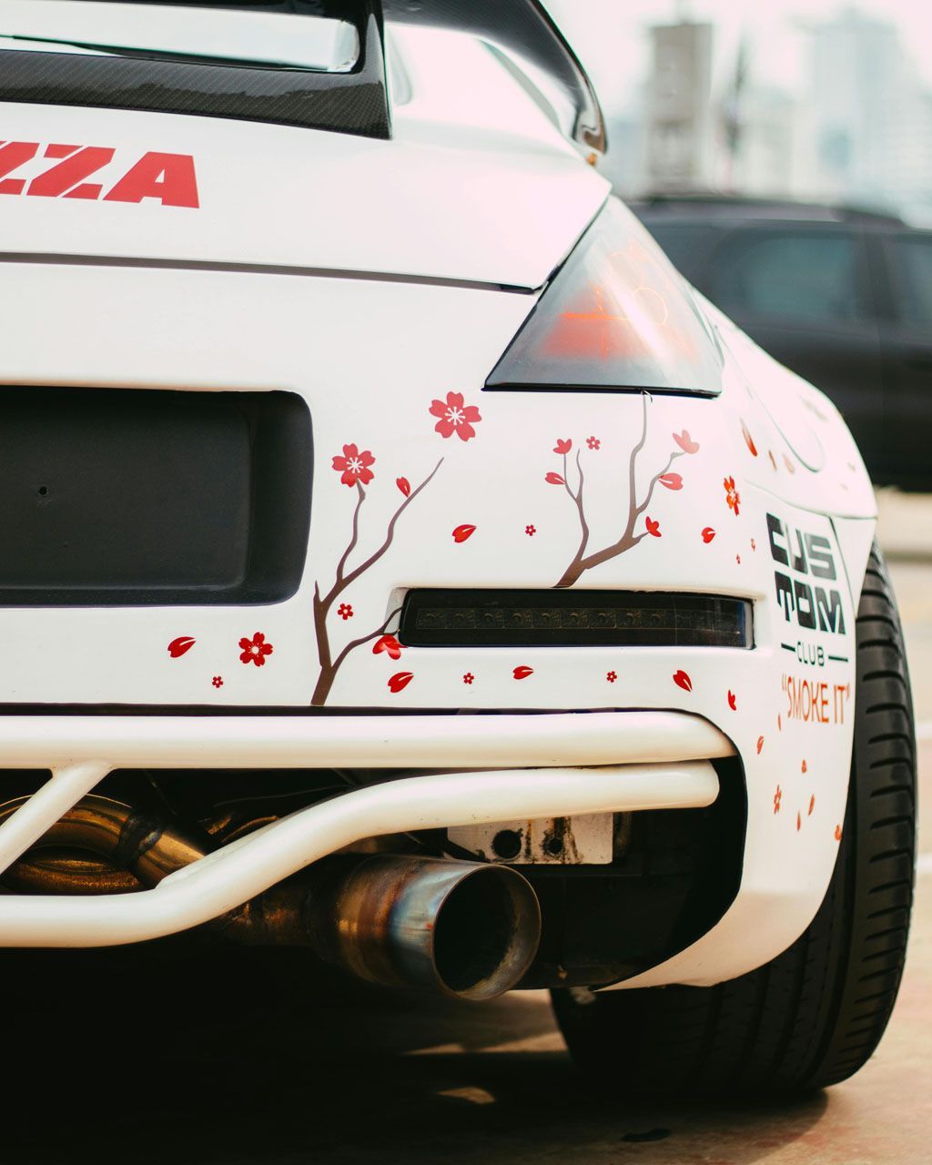 White car rear with cherry blossom decal, large exhaust pipe, and roll cage.