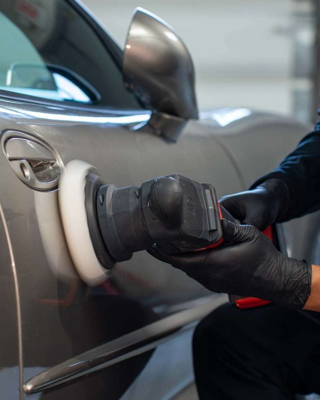 Person polishing a gray car with a power buffer, wearing black gloves.