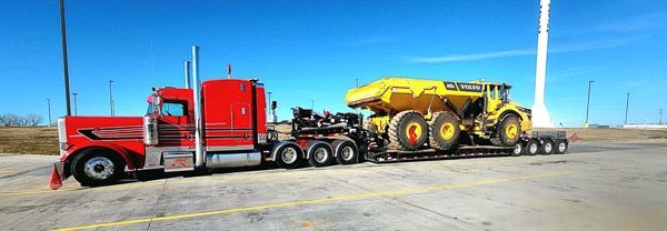 A red semi truck is carrying a yellow tractor on a trailer.