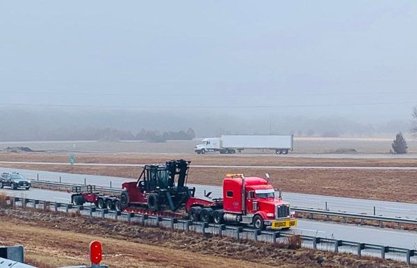 A red semi truck is driving down a highway carrying a forklift.