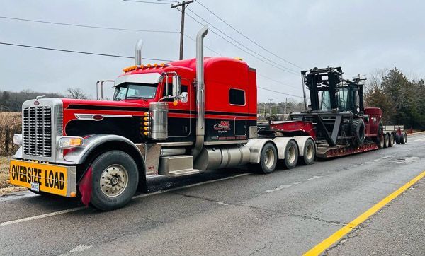 A red semi truck is carrying a forklift down a road.