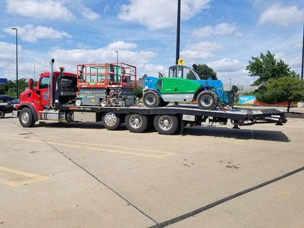 A red tow truck is carrying a green forklift on a flatbed trailer.