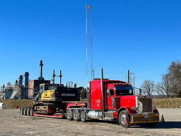 A red semi truck is carrying a yellow excavator on a trailer.