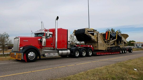 A red semi truck is carrying a large machine down a road.