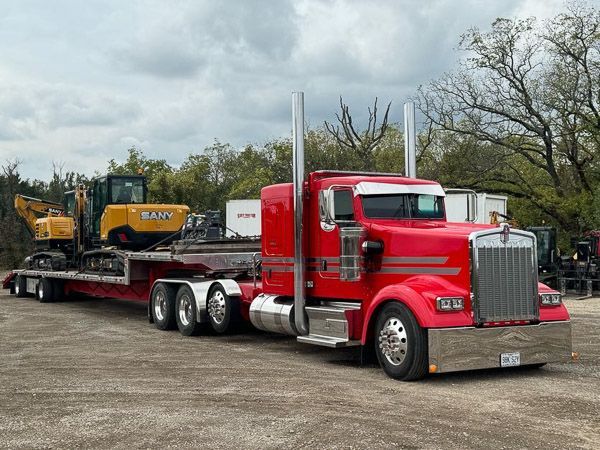 A red semi truck is carrying a yellow excavator on a trailer.