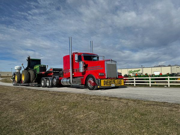 A red semi truck is pulling a green tractor down a road.
