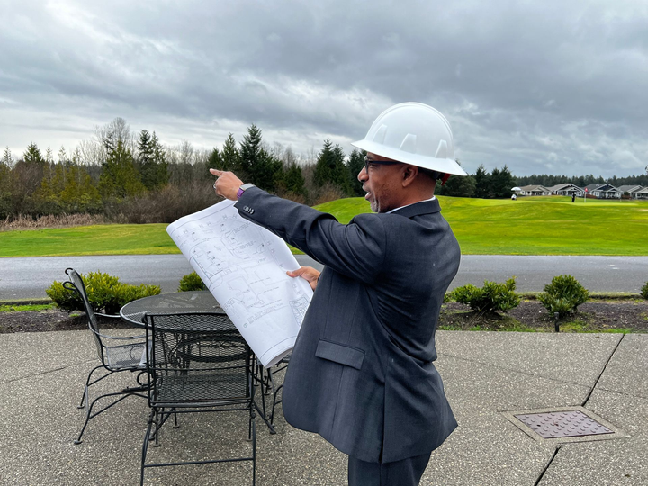 A man in a suit and hard hat is holding a blueprint and pointing at it.