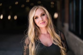 A woman with long blonde hair is wearing a black dress and standing in front of a building.