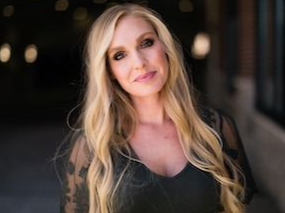 A woman with long blonde hair is wearing a black dress and standing in front of a building.