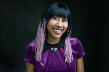 A woman with purple hair is wearing a purple shirt and smiling.