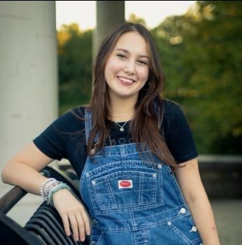 A woman in overalls is leaning against a car and smiling.