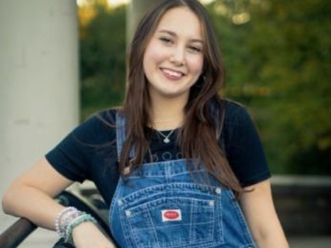 A woman in overalls is leaning against a car and smiling.