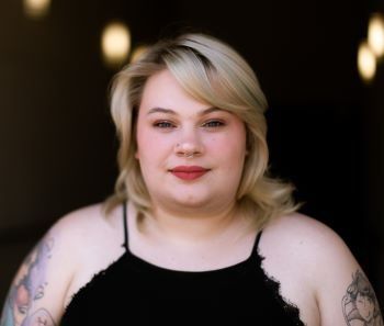 A woman with blonde hair and tattoos is wearing a black tank top.