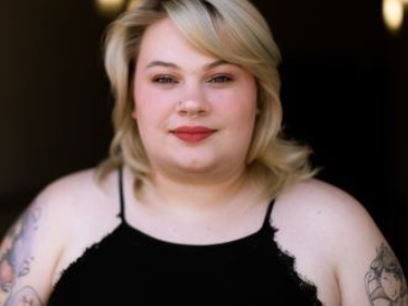 A woman with blonde hair and tattoos is wearing a black tank top.