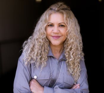 A woman with curly blonde hair is standing with her arms crossed and smiling.