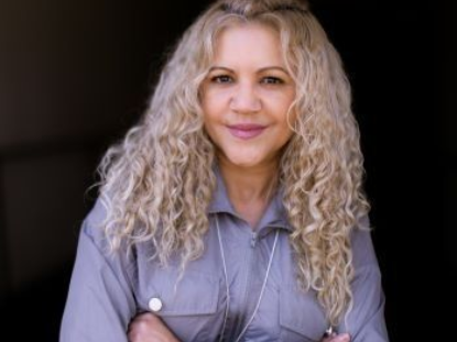 A woman with curly blonde hair is standing with her arms crossed and smiling.