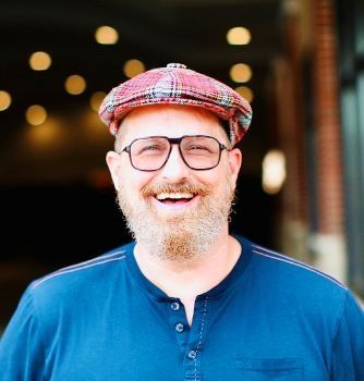 A man with a beard and glasses is wearing a hat and smiling.