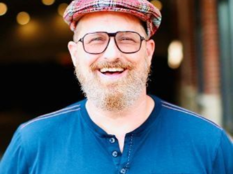 A man with a beard and glasses is wearing a hat and smiling.