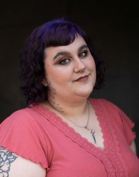 A woman with purple hair is wearing a pink shirt and a necklace.