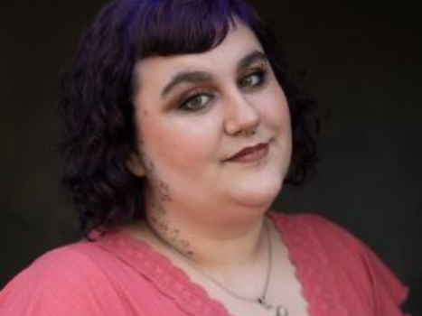 A woman with purple hair is wearing a pink shirt and a necklace.
