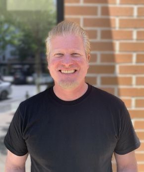 A man in a black shirt is smiling in front of a brick wall.