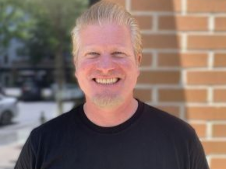 A man in a black shirt is smiling in front of a brick wall.