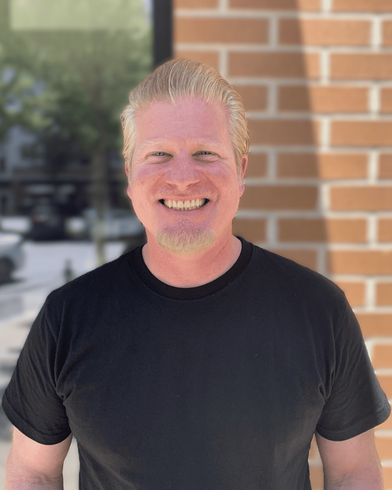 A man in a black shirt is smiling in front of a brick wall.
