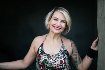 A woman with a tattoo on her arm is smiling and leaning against a wall.