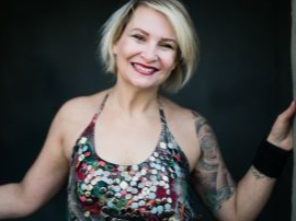 A woman with a tattoo on her arm is smiling and leaning against a wall.