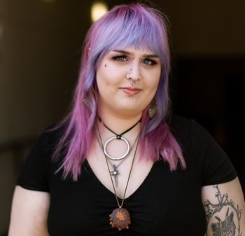 A woman with purple hair is wearing a black shirt and a necklace.