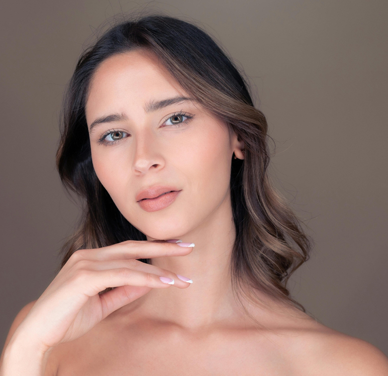 A person with shoulder-length wavy hair and natural makeup poses against a neutral background, hand near their chin.