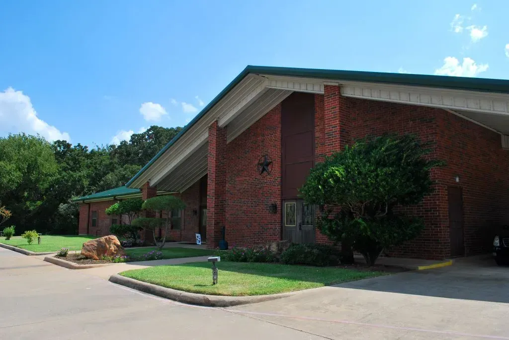 A low-angle exterior view of a brick building with a green gabled roof and a tree-lined lawn under a clear blue sky.