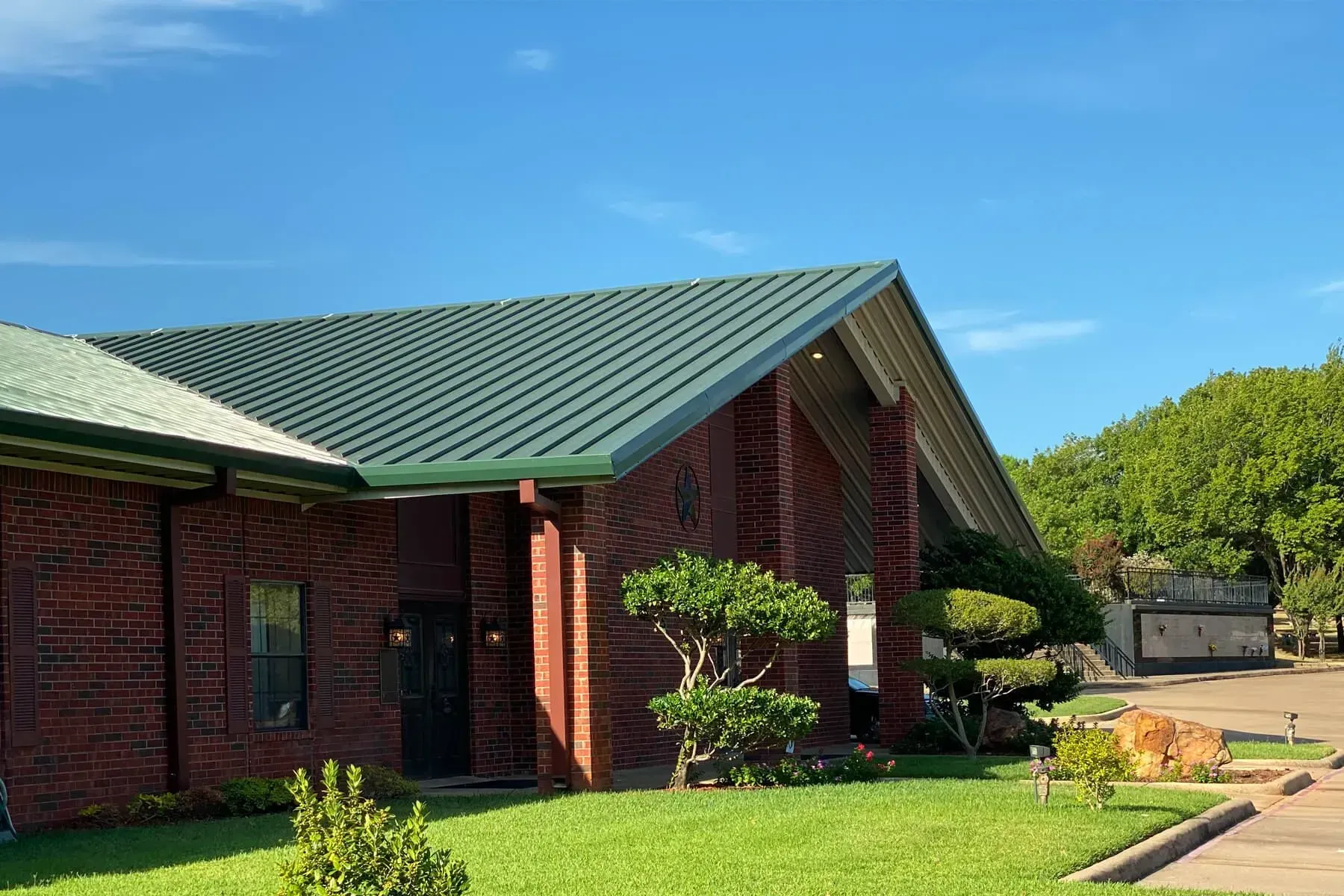 A brick building with a green metal roof under a bright blue sky, with manicured trees and a grassy lawn in front.