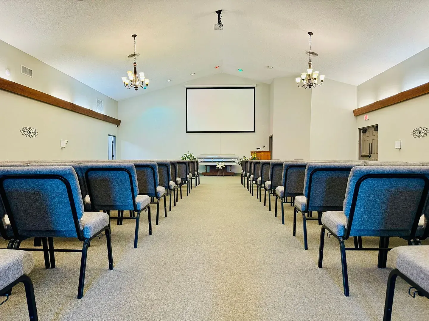 Interior of a church or hall with rows of blue upholstered chairs facing a stage featuring a white projector screen.