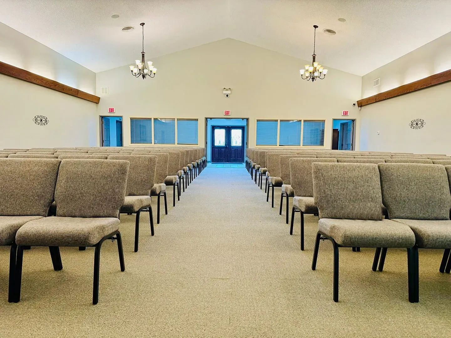 A symmetrical view of a church sanctuary with rows of grey cushioned chairs facing double doors under two chandeliers.
